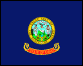 Image of the Idaho State Flag.  The Idaho State Flag measures five feet, six inches by four feet, four inches and is bordered by a gilt fringe two and one half inches wide. On a field of blue is positioned the Idaho State Seal, twenty-one inches in diameter, in colors, in the center of the blue field. On the seal, the women represent liberty, justice and equality; the man is a miner; the pictures on the shield represent the main industries of forestry, farming and mining; the cornucopias, or horns of plenty are symbols of abundance; the elk's head represents wildlife; and the motto in Latin above the elk's head -  Esto perpetua - means 'Let it be perpetual'. The words 'State of Idaho' are embroidered in with block letters, two inches in height on a red band three inches in width by twenty-nine inches in length, the band being in gold and placed about eight and one-half inches from the lower border of fringe and parallel with the same. The flag was adopted in 1907.