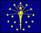Image of the Indiana State Flag.  The field of the flag is blue with nineteen stars and a flaming torch in gold or buff. Thirteen stars are arranged in an outer circle, representing the original thirteen states; five stars are arranged in a half circle below the torch and inside the outer circle of stars, representing the states admitted prior to Indiana; and the nineteenth star, appreciably larger than the others and representing Indiana is placed above the flame of the torch. The outer circle of stars are arranged so that one star appears directly in the middle at the top of the circle, and the word 'Indiana' is placed in a half circle over and above the star representing Indiana and midway between it and the star in the center above it. The torch stands for liberty and enlightenment. Six rays are shown radiating from the torch to the three stars on each side of the star in the upper center of the circle, and are meant to sybolize the expansive nature of those two concepts as well as mean that freedom and knowledge are available to everyone. The flag was adopted in 1917.