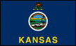 Image of the Kansas State Flag.  The flag displays the Kansas State Seal centered on a blue field. Above the seal is the state crest, a sunflower resting on a twisted blue and gold bar. Below the seal in gold block lettering is the name of the state, 'KANSAS'. The State Crest consists of a twisted blue and gold bar representing the Louisiana Purchase, from which Kansas was carved. The state flower, the Sunflower, is depicted as if 'torn from its stalk.' Some say that the 'open frankness of the Sunflower is indicative of the fearlessness with which Kansas meets her problems and solves them.' The State Seal centered on the flag tells, eloquently, the story of Kansas. The thirty-four stars clustered at the top of the seal identify Kansas as the 34th state to be accepted into the Union. Above the stars is printed the state motto, 'Ad Astra per Astera', Latin for 'To the Stars through Difficulties,' meaning that the people of Kansas do not let difficulties stop them from doing their best. The rolling hills around Fort Riley are reflected in the seal. The Indians hunting American Bison, the prairie schooners heading west and the oxen mirror the expanding frontier. The farmer plowing his field before his log cabin at sunrise represent agricultural assets. The steamboat navigating the Kansas River, delivering supplies to Fort Riley, represent commerce within the state. The flag was adopted in 1927.