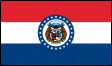Image of the Missouri State Flag.  The flag consists of three horizontal red, white and blue stripes. These stripes represent valor, purity and vigilance and justice. A circle is centered on the flag surrounded by a band of blue enclosing the Missouri Coat of Arms on a white background. The blue band displays 24 white five-pointed stars representing Missouri as the 24th state. The shield of the Missouri Coat of Arms shows, on the right, a Bald Eagle grasping the olive branches of peace and the arrows of war in its talons. This represents the strength and powers of the Federal Government. On the left side of the shield, the state side, is a grizzly bear and a crescent moon. The grizzly bear symbolizes the strength and bravery of the citizens of the state. The crescent moon sybolizes the state of Missouri at the time of its induction into the union; a state with a small population and wealth and huge potential. The crescent moon also symbolizes the 'second son.' Missouri was the second state to be carved from the territory aquired with the Louisiana Purchase. The shield is encircled by a belt inscribed 'United we stand, divided we fall' indicating the advantage of the union of the United States. Two more grizzly bears, one on each side of the shield, echo the bravery and strength of the state's citizens. They are standing on a scroll displaying the Missouri State Motto, 'Salus Populi Suprema Lex Esto' (Let the welfare of the people be the supreme law). Below the scroll are the Roman Numerals for 1820, the year that Missouri became a member of the United States. Above the shield a helmut is depicted, representing Missouri as a sovereign state. A large star surrounded by 23 smaller stars signifies Missouri's status as the 24th state. A cloud around the large star represents the difficulties that Missouri endured on its way to statehood. The flag was adopted in 1913.
