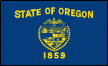 Image of the Oregon State Flag.  The flag is colored navy blue with gold lettering and symbols. Certain occasions demand a more formal version of the flag and so a dress or parade version of the flag is fringed in gold. The flag face displays the words 'STATE OF OREGON' above a gold shield surrounded by thirty-three stars. The stars represent Oregon as the thirty-third state to be admitted to the union. Below the shield, part of the state seal, is the date '1859' in gold numerals. This is the year that Oregon was admitted to the union. Depicted on the shield, is a blazing sun setting over the Pacific Ocean, mountains, forests and a conestoga wagon all representing the natural resources and splendor of Oregon and the early settlers to the territory. A plow, a sheaf of wheat and a pickax represent the early industry of the state; agriculture and mining. Two ships are shown; a British Man-of-War and an American trade vessel. The departing British ship and arriving American ship are said to represent that claim to the land was laid by both Great Britain and The United States at the same time. It is also said that the tack of the two ships represents the ascendence of American power in the western hemisphere. Another interpretation has the departure and arrival as representative of trade or commerce. This explanation would seem to ignore that the British vessel is a Man-of-War. The crest of the shield is the American eagle. Also displayed is a banner with the words 'The Union.' The eagle and the banner express support for 'The Union' that Oregon joined in 1859. The Oregon State Flag is the only state flag with a different design on each side. With a nickname of 'The Beaver State' one might easily guess the depiction on the obverse side of the flag. Indeed, the Oregon State Animal makes a strong showing on the Oregon State Flag. The flag was adopted in 1925.