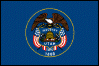 Image of the Utah State Flag.  The flag displays the Utah State Seal centered on a blue background and circled by a thin gold line. At the center of the Great Seal of the state of Utah is a shield with a beehive, the state emblem, with a sego lily growing on either side. The sego lily stands for peace. Atop the beehive is the word 'Industry', the state motto. 'Industry' means steady effort. Even prior to Utah gaining statehood in 1896, the image of the beehive and the term Industry were unofficial symbols of the region. The 1847 provisional state of Deseret, which was to become Utah, adopted the beehive as its official emblem, and the symbol carried through as Utah became a part of the Union. The qualities of the beehive (industry, perseverance, thrift, stability, and self-reliance) were all virtues respected by the region's settlers. On both sides of the shield are American flags, showing that Utah supports the United States. Atop the shield is an American eagle, which stands for protection in peace and war. Under the shield the date 1847 represents the year that Brigham Young led a group of people to the Salt Lake Valley to reestablish the Church of Jesus Christ of Latter Day Saints, also known as the Mormons. At the bottom of the seal the date 1896 represents the year that Utah gained admission to the United States. The flag was adopted in 1913.