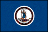 Image of the Virginia State Flag.  The flag has a deep blue field with a circular white center that contains the obverse side of the Great Seal of Virginia, with 'Virginia' at the top of the seal and the Latin motto 'Sic Semper Tyrannis' - 'Thus Always to Tyrants' at the bottom. Adopted in 1776, when Virginia and the other colonies were fighting for independence from England, it shows two figures acting out the meaning of the motto. Both are dressed as warriors. It depicts the Roman goddess Virtus representing the spirit of the Commonwealth. She is dressed as an Amazon, a sheathed sword in one hand, and a spear in the other, and one foot on the form of Tyranny, who is pictured with a broken chain in his left hand, a scourge in his right, and his fallen crown nearby, implying struggle that has ended in complete victory. A border of Virginia creeper, in red and green colors, encircles the design of the seal, and a white silk fringe adorns the edge of the flag farthest from the flag staff. The flag was adopted in 1861.