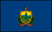 Image of the Vermont State Flag.  The flag displays a design derived from a painting made for the Vermont State Coat of Arms on a blue field. It is based on the state seal that was adopted in 1779. A gold shield contains a tall pine tree, a cow and sheaves of wheat. The Green Mountains are in the distance. Pine boughs extend around the shield. The name 'Vermont' and the state motto 'Freedom and Unity' are displayed on a crimson banner below the shield. Above the shield is a stag's head. The flag was adopted in 1923.